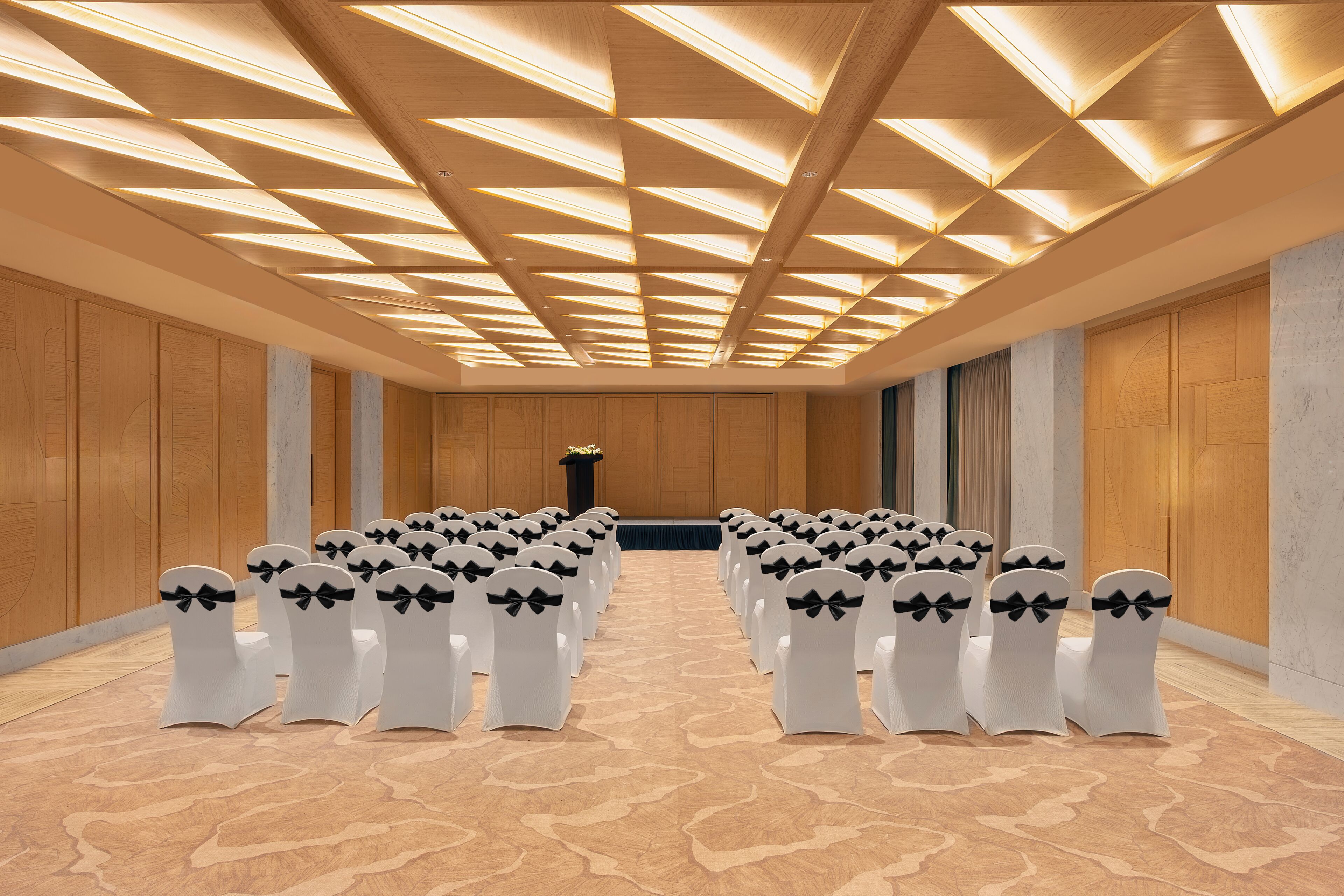 A conference room with rows of white chairs, a patterned carpet, wooden panel walls, and a ceiling with geometric lighting.