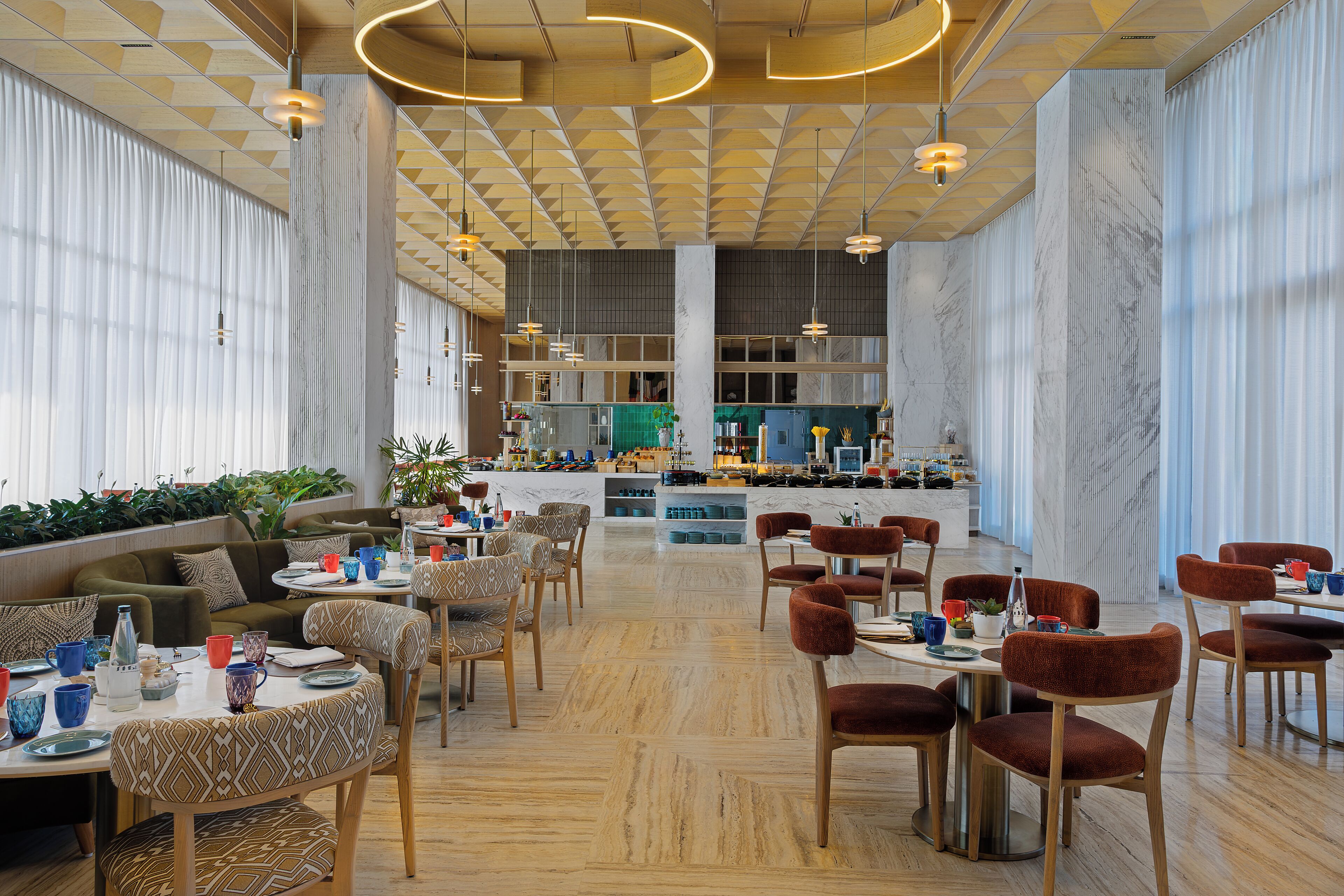 A spacious dining area with multiple tables and chairs, a kitchen in the background, and large windows with curtains.