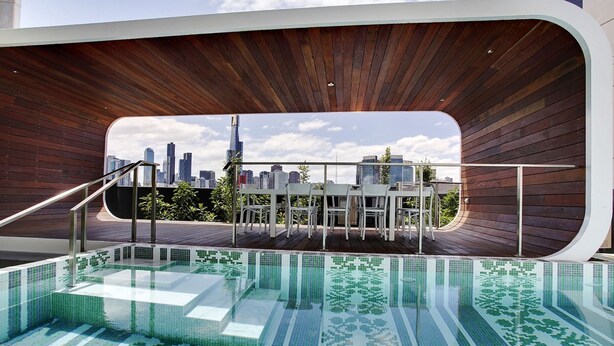 A modern outdoor pool with a city skyline view, wooden deck, and a glass-enclosed seating area. - Chic City Escape With Pool, Gym & Parking (Melbourne)