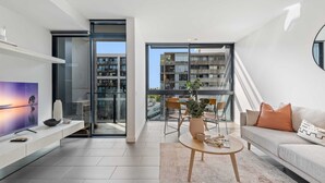 A modern living room with a flat-screen TV, a sofa, a coffee table, and a dining area with chairs and a table. - Chic City Escape With Pool, Gym & Parking (Melbourne)