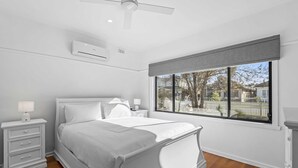 A bedroom with a bed, a nightstand, a lamp, a window with a view, and an air conditioning unit. - Spacious 3-bed With Outdoor Dining Near Airport (Melbourne)