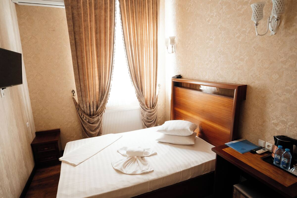 Une chambre d’hôtel avec un lit, une table de chevet avec une lampe, un téléviseur fixé au mur et une fenêtre avec des rideaux.