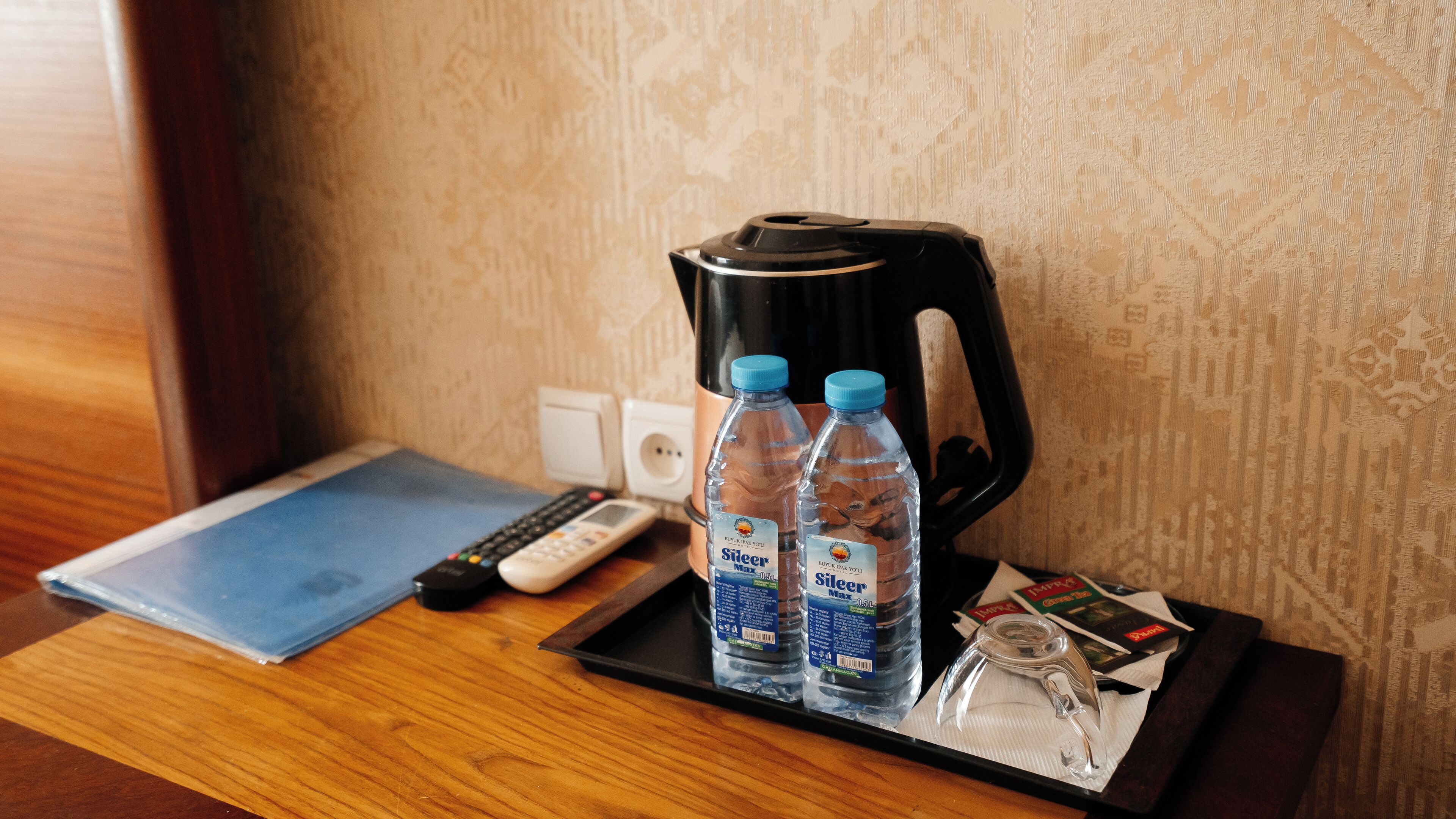 Ein Hotelzimmer mit Holztisch, einer schwarzen Tablett mit zwei Wasserflaschen, einer Fernbedienung, einem Wasserkocher und einem Glas.