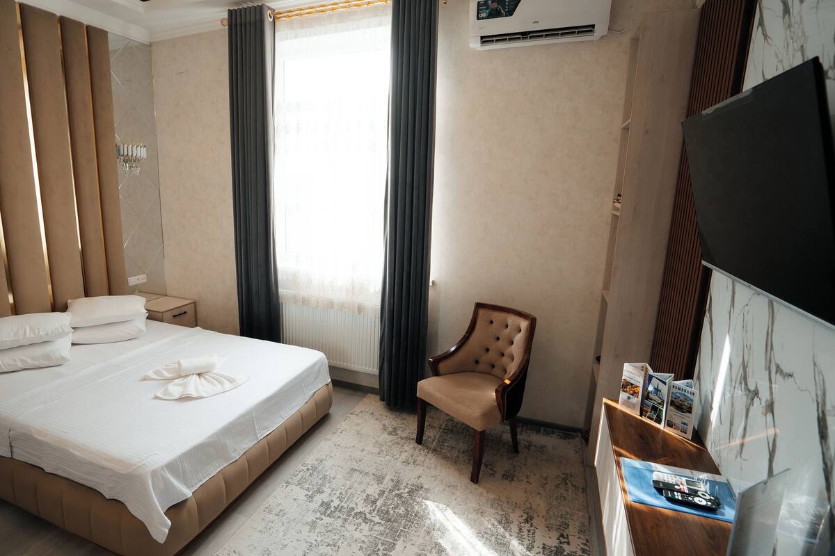 Une chambre d’hôtel avec un grand lit, une table de chevet, une chaise, une télévision et une fenêtre avec des rideaux.