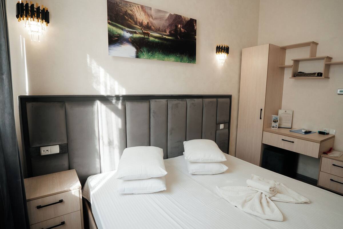Une chambre d’hôtel avec un grand lit, une table de chevet, un bureau et une chaise. Il y a un tableau au mur et une fenêtre avec des rideaux.