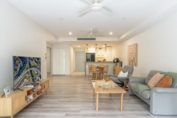 A modern living room with a sofa, coffee table, and TV. There is a dining area with chairs and a table. The kitchen is visible in the background. - Best Location 2-bed With Gym, Pool 7-min From CBD (Brisbane)