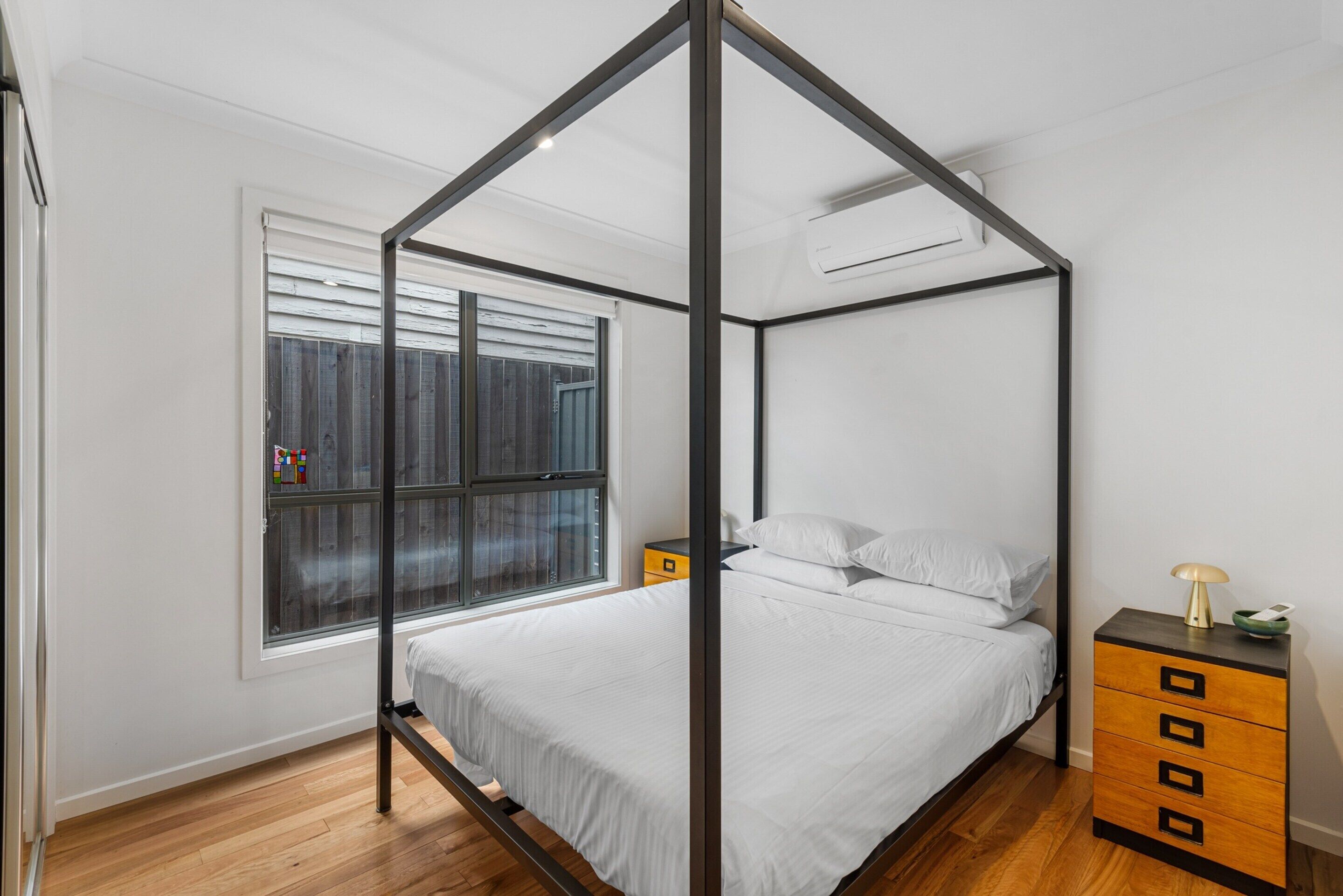 Ein Schlafzimmer mit Himmelbett, Holzboden, einem Fenster mit Jalousien, einem Nachttisch mit Lampe und einer kleinen Kommode.