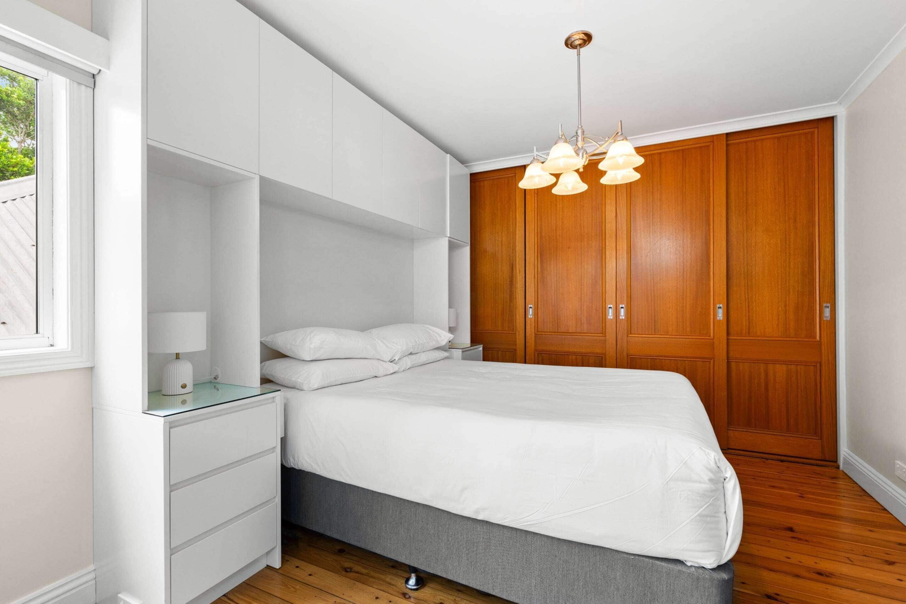 A bedroom with a bed, white nightstands, a glass-top dresser, a wooden wardrobe, and a hanging light fixture.
