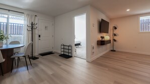 A modern living room with a wooden floor, a dining area with a table and chairs, a coat rack, a TV mounted on the wall, and a large window with a view of the outside. - Nordic Suite W | Private, Fenced Yard, Outdoor Patio w/ Fire-Table, A/C, Laundry (Lethbridge)