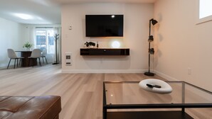 A modern living room with a flat-screen TV mounted on the wall, a glass coffee table, a brown leather bench, and a dining area in the background. - Nordic Suite W | Private, Fenced Yard, Outdoor Patio w/ Fire-Table, A/C, Laundry (Lethbridge)