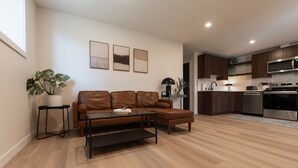 A modern living room with a brown leather sofa, a coffee table, and a side table with a plant. There is a kitchen area with dark cabinets, a microwave, and a dishwasher. - Nordic Suite W | Private, Fenced Yard, Outdoor Patio w/ Fire-Table, A/C, Laundry (Lethbridge)
