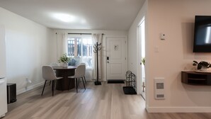 A modern living room with a dining area, a flat-screen TV mounted on the wall, and a large window with curtains. - Nordic Suite W | Private, Fenced Yard, Outdoor Patio w/ Fire-Table, A/C, Laundry (Lethbridge)