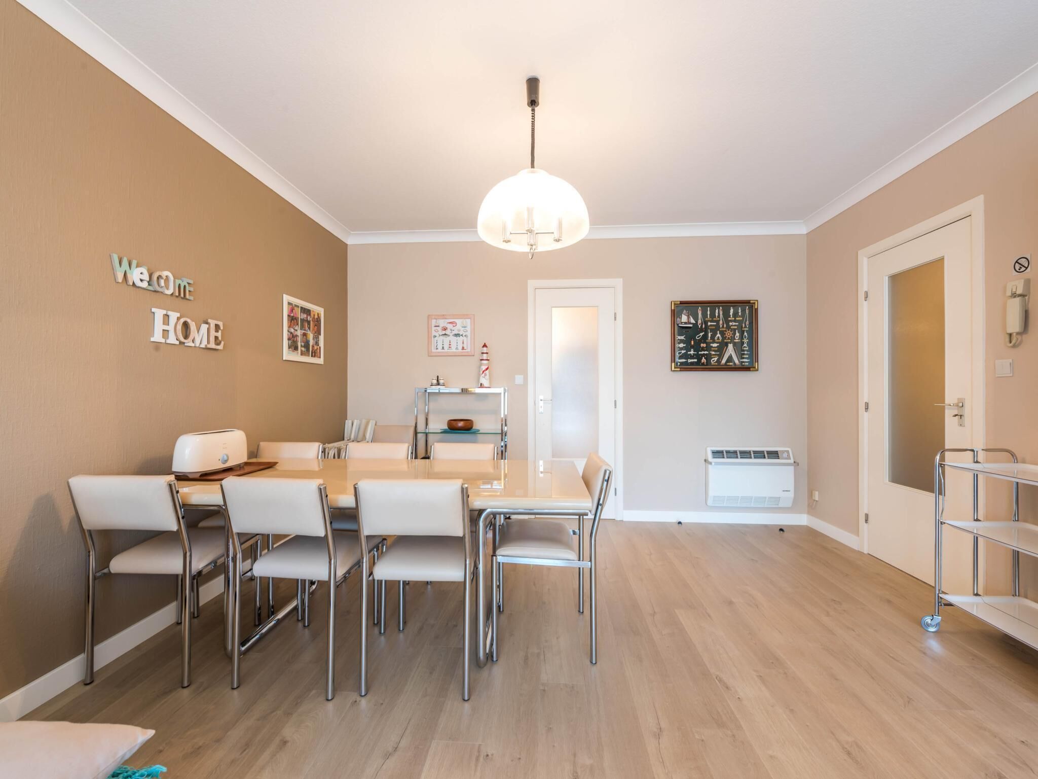 A modern dining area with a white table and chairs, a chandelier, and a "Welcome Home" sign on the wall.