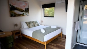 Una cama individual con estructura de madera, una mesita de noche, una silla, un cuadro en la pared y una ventana con vistas a zonas verdes.