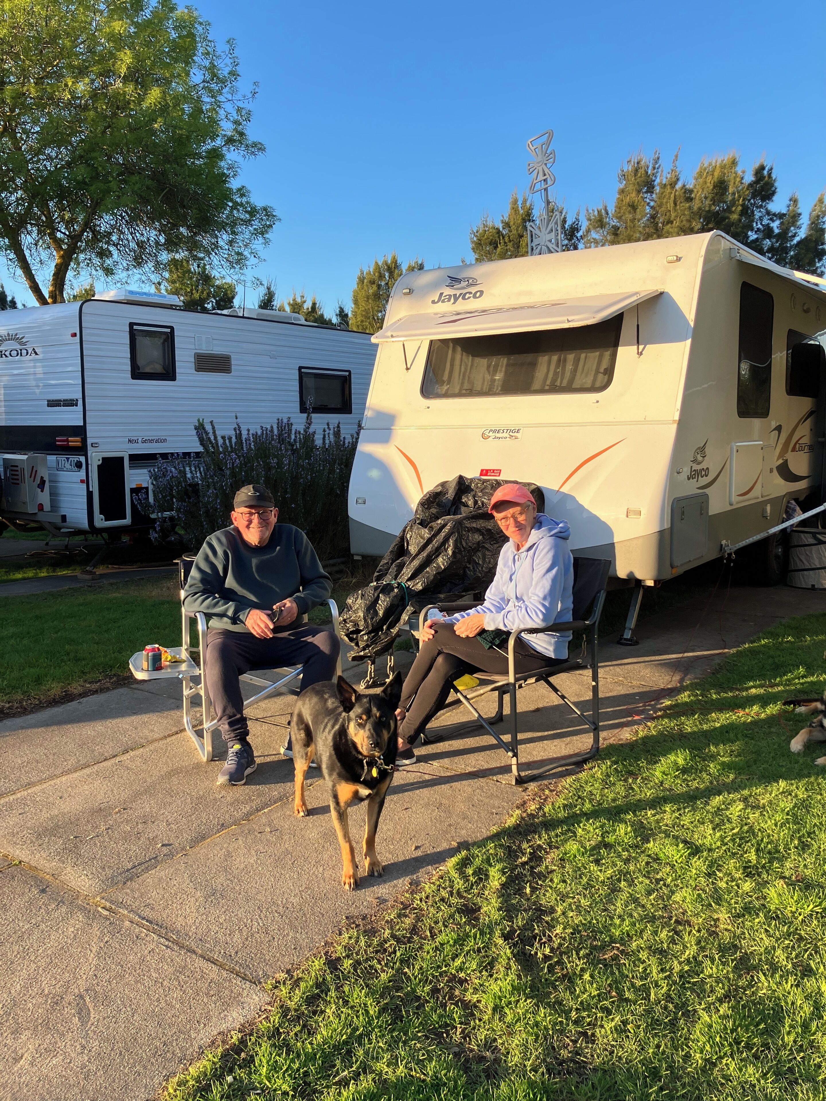 Två personer sitter på stolar med en hund emellan sig. I förgrunden syns en vit husbil med texten ”Jayco”, och längre bak syns en husbil av märket ”KODA”.