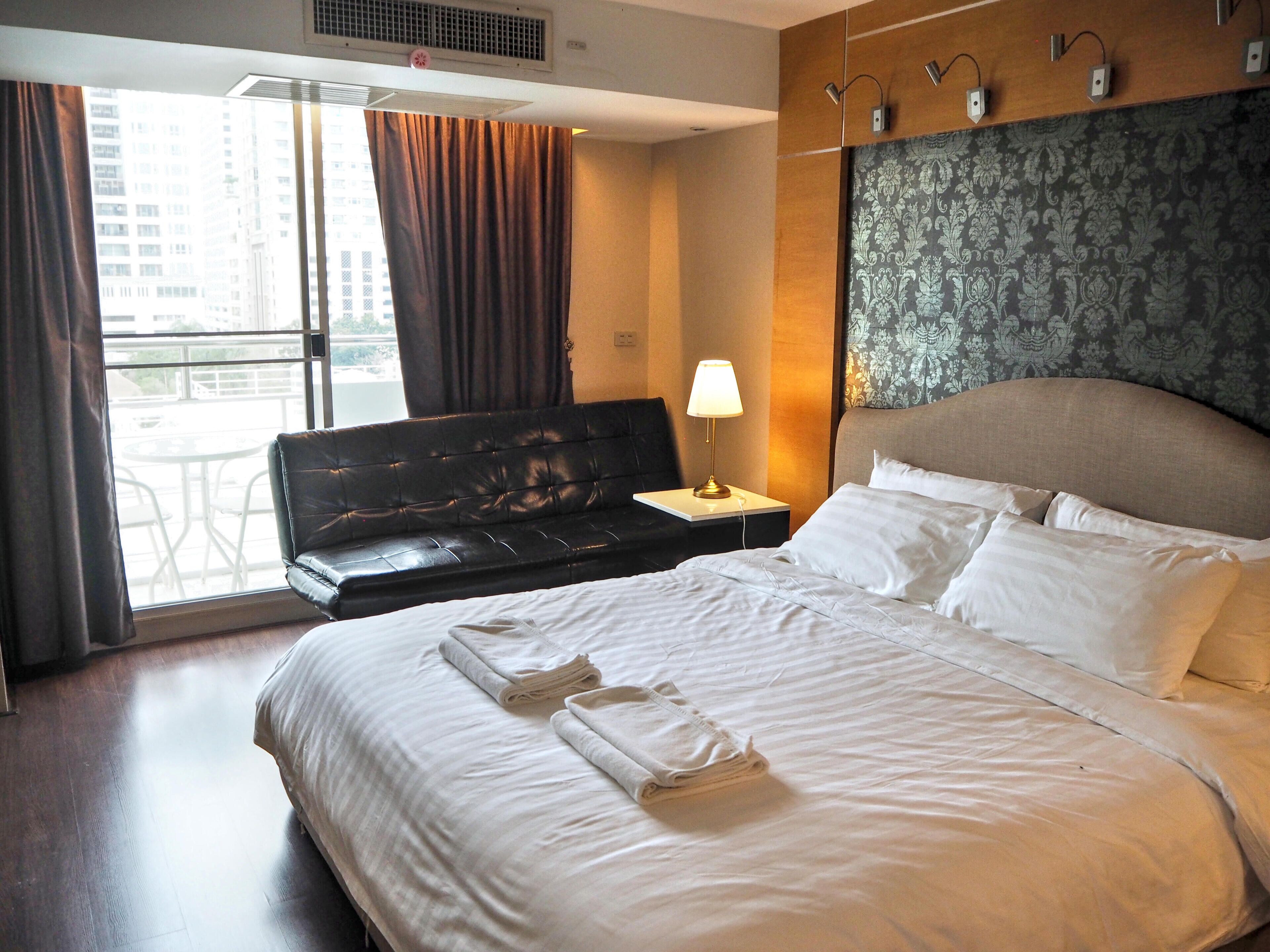 Habitación de hotel con cama, mesita de noche, lámpara, ventana con cortinas y vista a edificios.