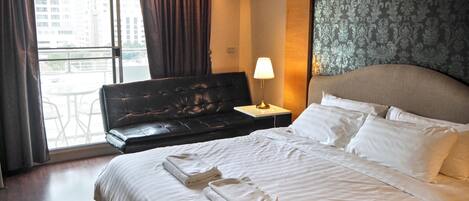 Habitación de hotel con cama, mesita de noche, lámpara, ventana con cortinas y vista a edificios.