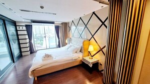 A hotel room with a large bed, a bedside table with a lamp, a window with curtains, and a view of the city. - Harmony Living Condominium (Bangkok)