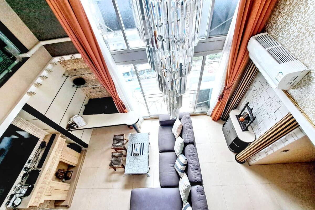 A modern living room with a sofa, a coffee table, and a TV. There is a staircase leading to an upper level, and a window with curtains.