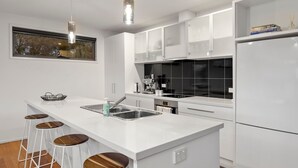 A modern kitchen with white cabinetry, a central island with stools, a built-in oven, and a coffee machine. - Idyllic Aireys Retreat - where ocean & hills meet (Aireys Inlet)