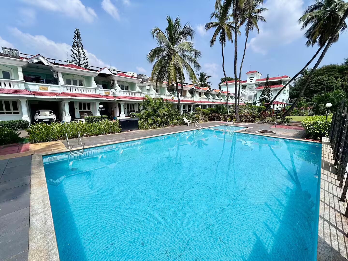 A swimming pool with a clear blue water surface, surrounded by a poolside area with lounge chairs and a building with a red roof in the background.