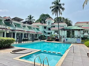 A swimming pool with a surrounding patio area, a covered seating area, and a multi-story building with balconies.