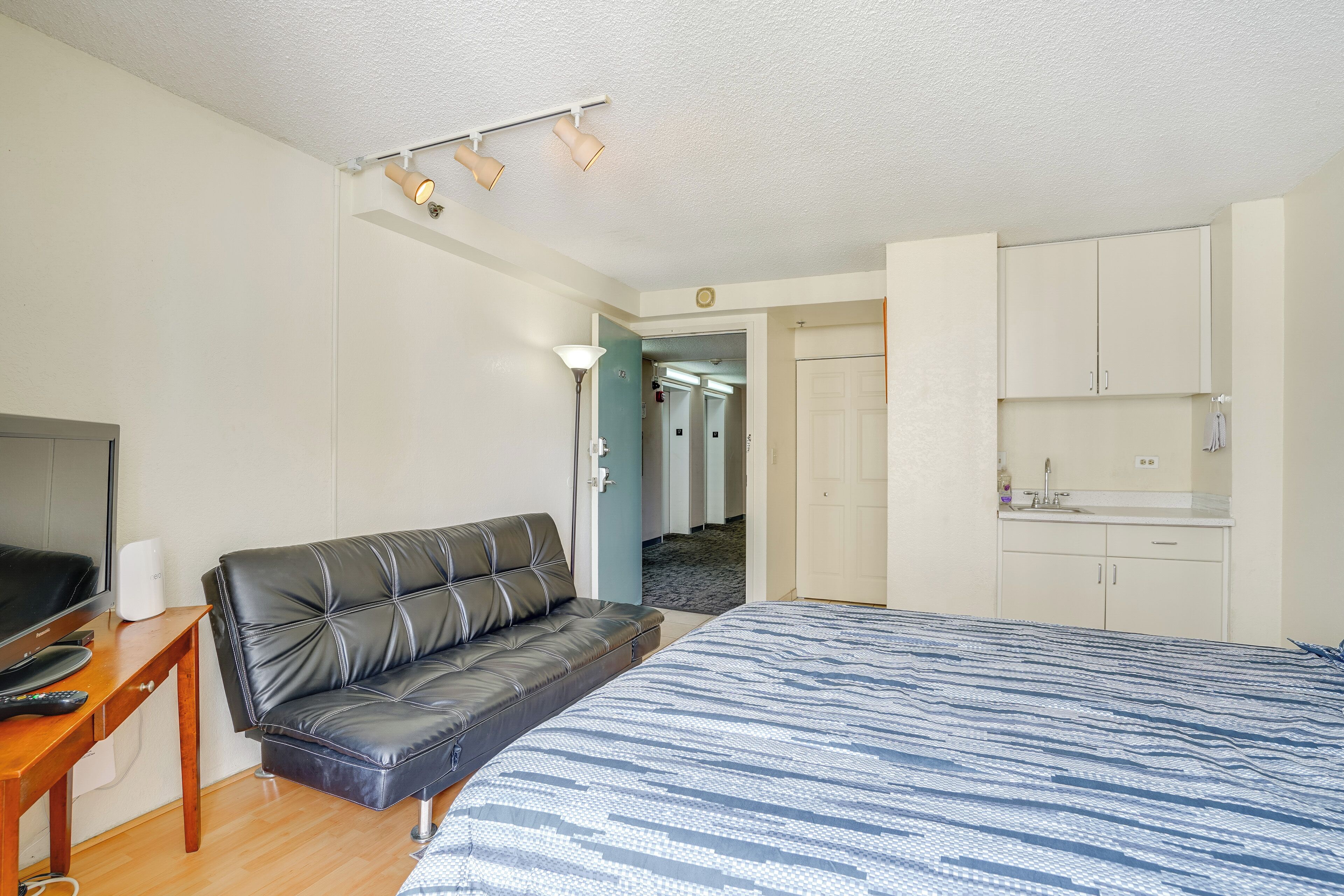 A hotel room with a bed, a leather sofa, a TV, a desk, and a kitchenette.