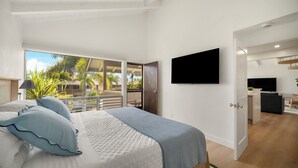 Et moderne soveværelse med en stor seng, et fjernsyn ophængt på væggen og en balkon med udsigt til palmer.
