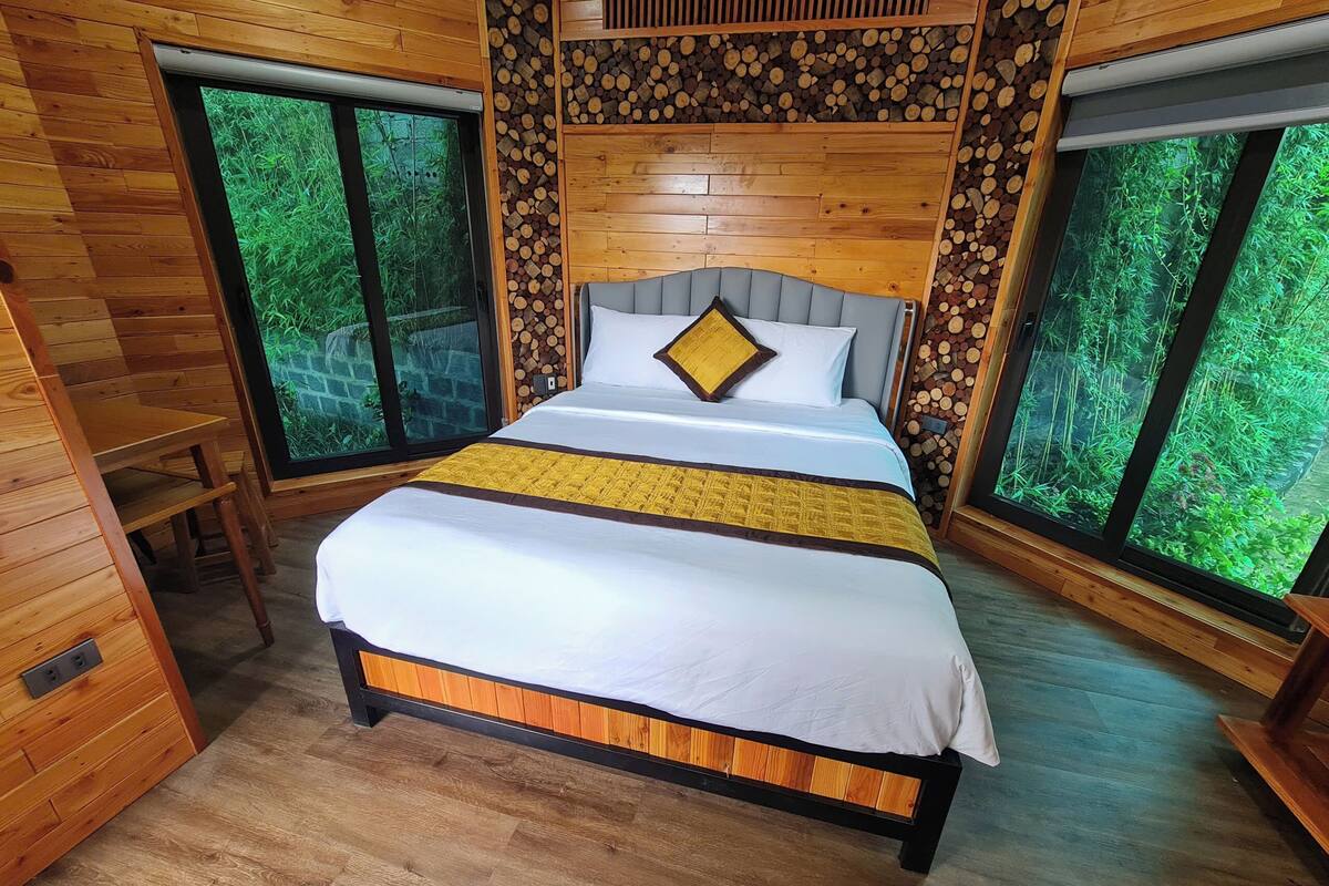 Una habitación de madera con una cama, un banco y una ventana que ofrece vistas al verdor.