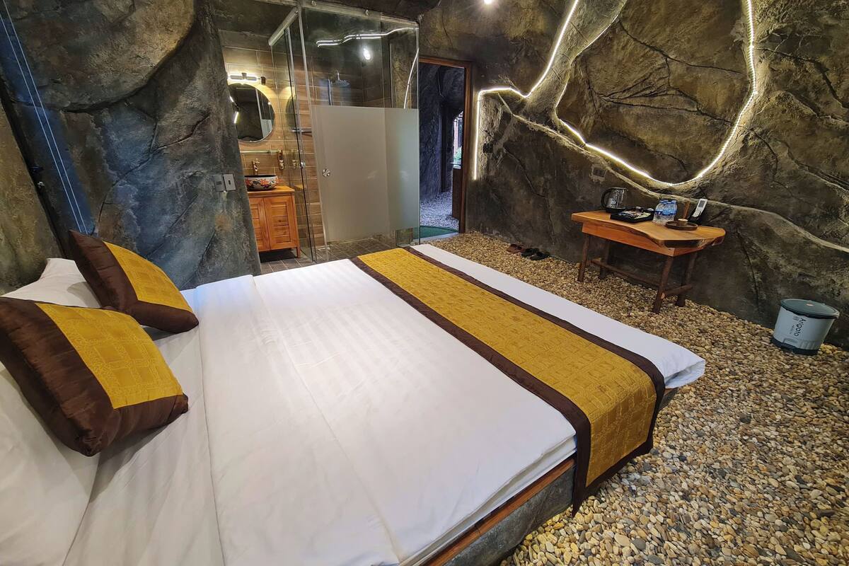 Un dormitorio con cama, una mesita de noche de madera, un espejo y un baño visible a través de una puerta de vidrio.