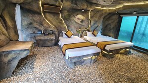 Ein Zimmer mit einer einzigartigen, strukturreichen Decke, zwei Betten mit weißer Bettwäsche und goldenen Akzenten, ein kleiner Tisch mit Wasserkocher, Ventilator und einem Fenster mit Blick auf den Wald.