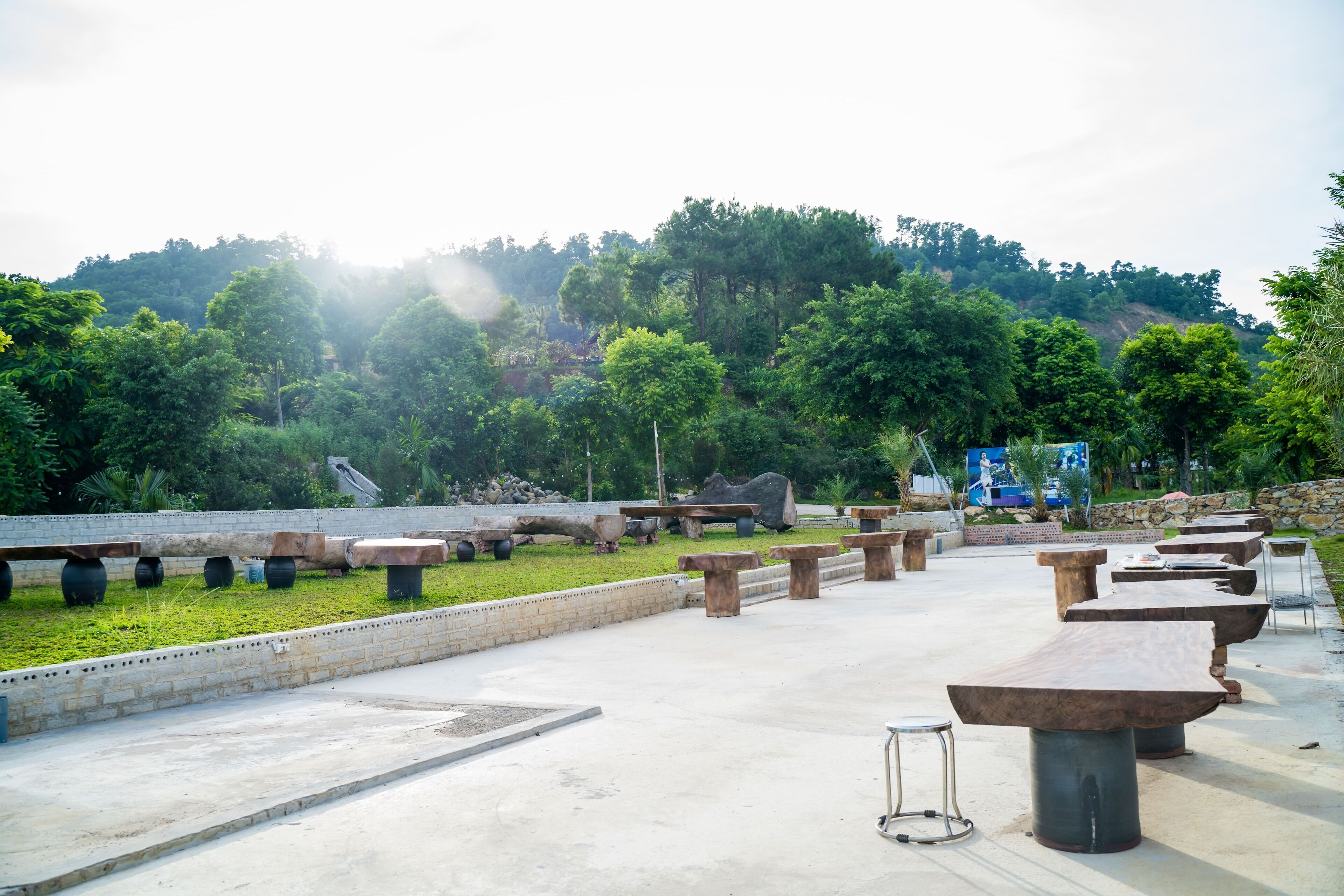 An outdoor seating area with stone tables and benches, surrounded by greenery and a hilly landscape.