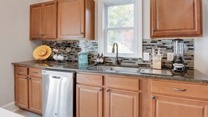 A kitchen with wooden cabinets, a stainless steel dishwasher, a window with a view of trees, and a tiled backsplash.