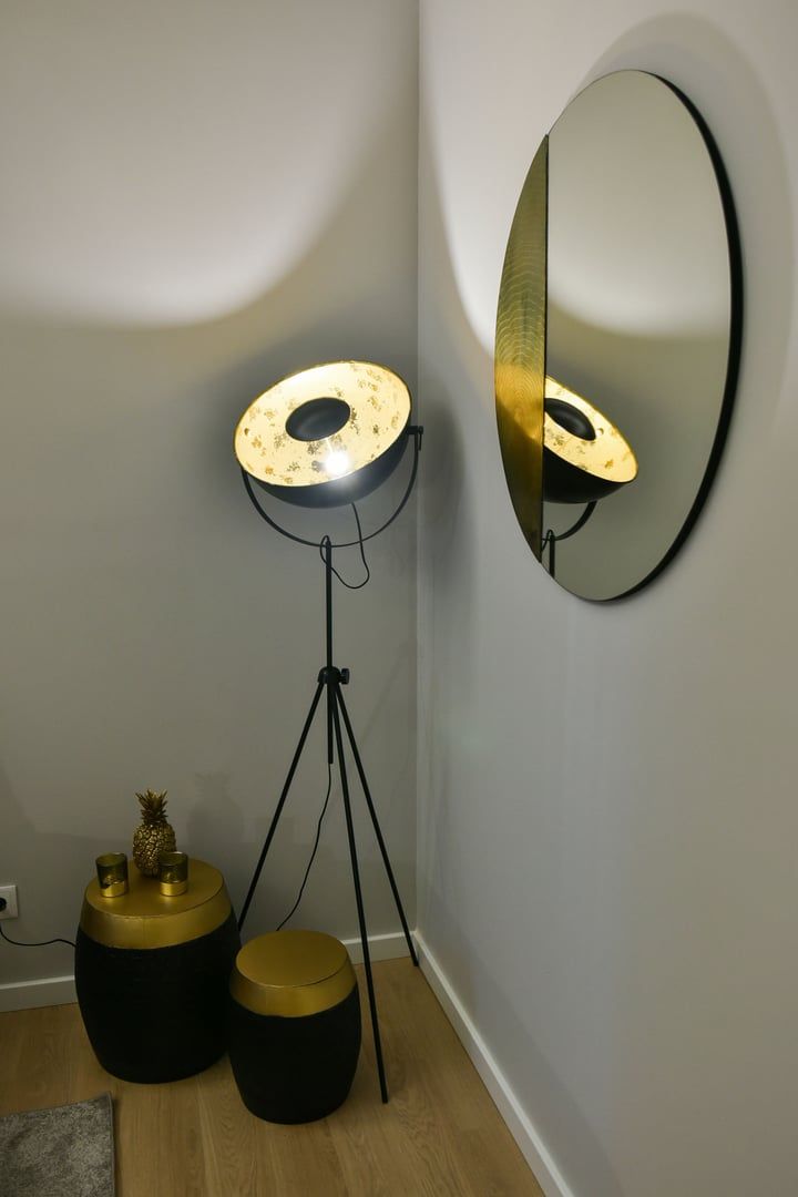 Een hoek met een vloerlamp, een ronde spiegel en twee zwarte bijzettafels met goudkleurige accenten.