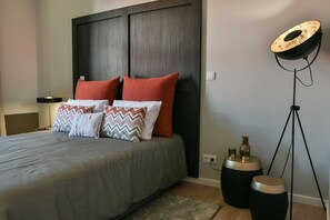 Een moderne slaapkamer met een grijze spreidek, twee rode kussens en decoratieve kussens met ruitmotieven.