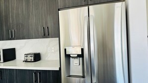 Un refrigerador de acero inoxidable con dispensador de agua, un microondas y un tostador sobre una encimera blanca.