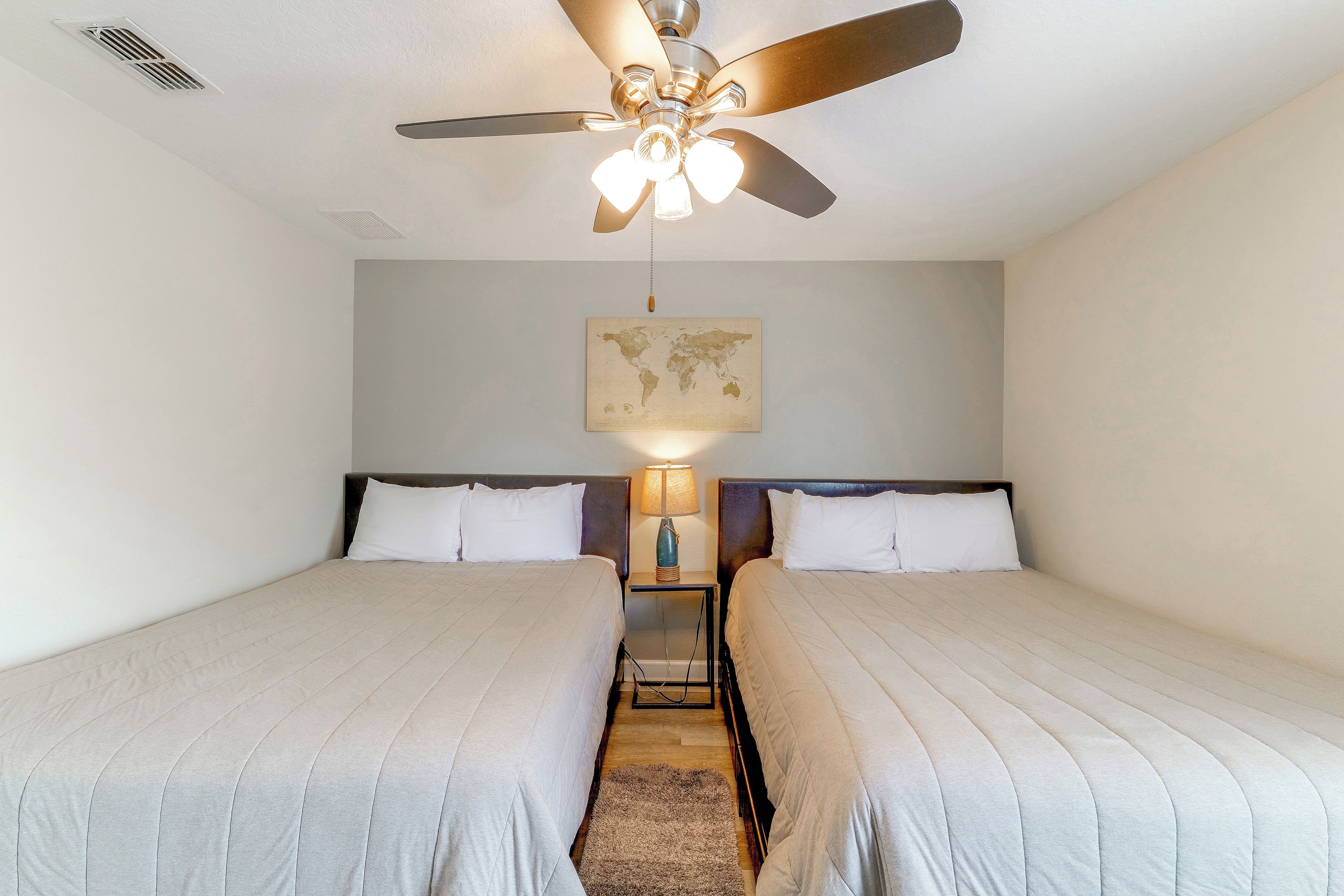 A hotel room with two single beds, a ceiling fan, a world map artwork, and a nightstand with a lamp.