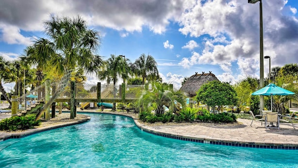 A clear blue pool with a wooden bridge, surrounded by palm trees and a thatched-roof structure. - ChampionsGate Resort Perks! Fun-Filled Family Home (Davenport)