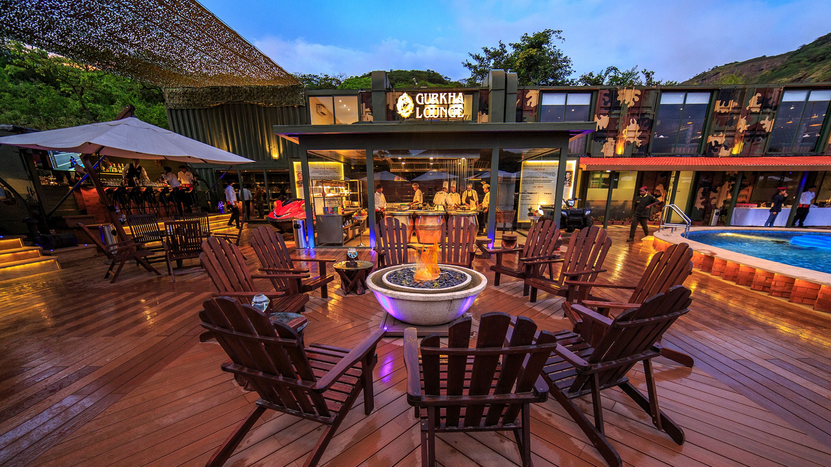 A cozy outdoor seating area with wooden chairs and a central fire pit, surrounded by a pool and a building with a sign reading "GURKHA LOUNGE".