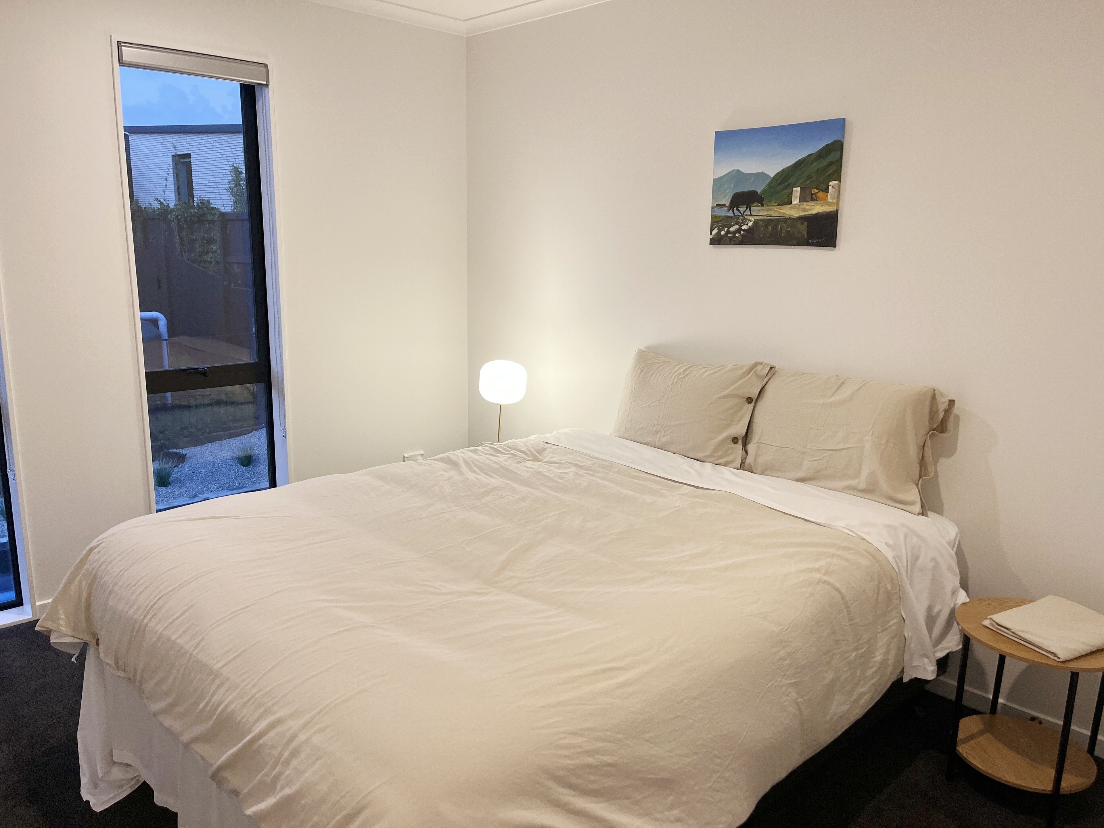 Une chambre avec un grand lit, une table de chevet avec une lampe, un tableau au mur et une vue sur l’extérieur par une porte.