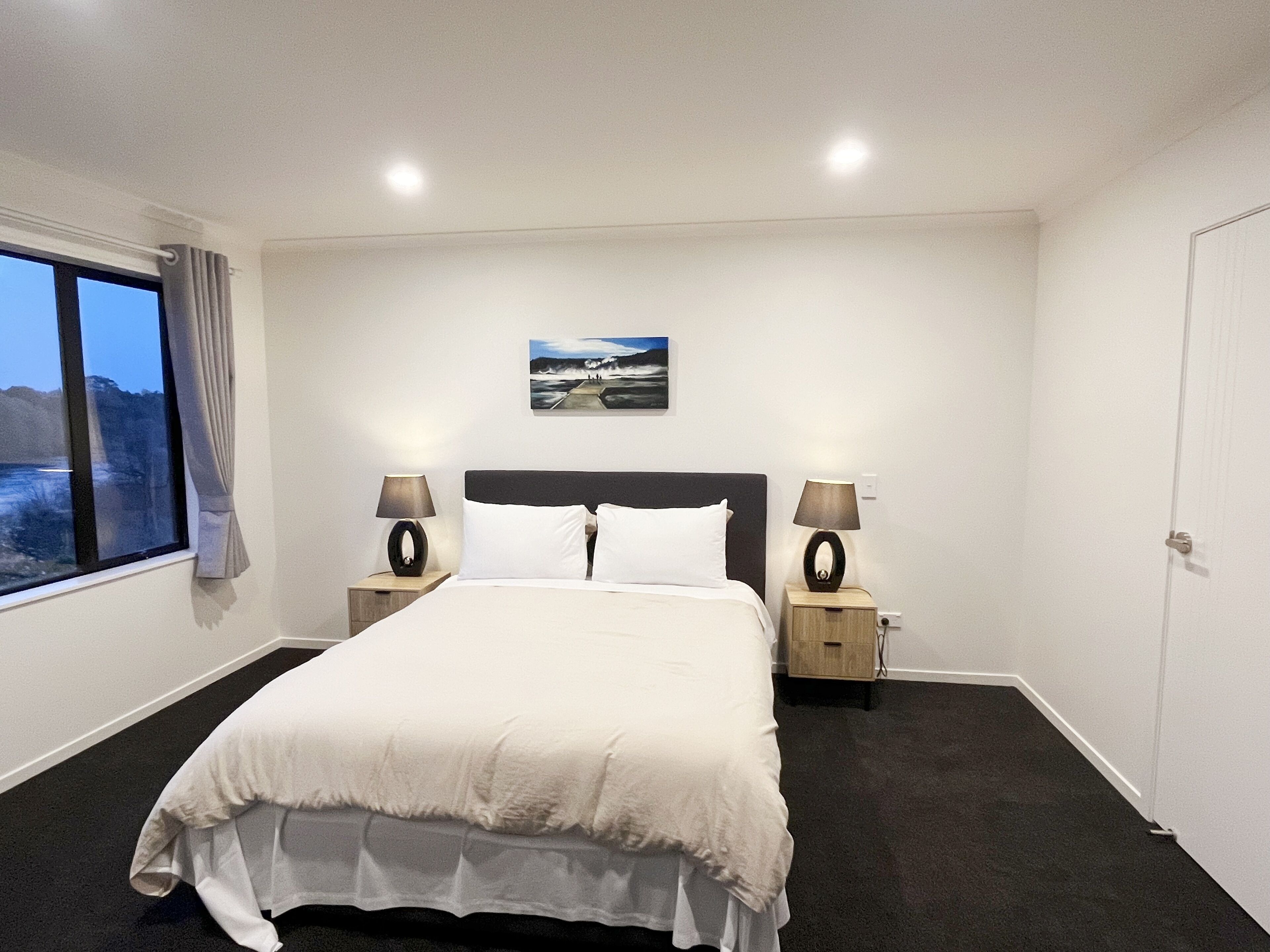 A hotel room with a large bed, two bedside tables with lamps, a window with curtains, and a painting on the wall.