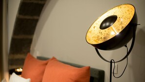 A modern lamp with a unique design, placed on a stand in a room with a bed and orange pillows. - GuestReady - Casa das Arcadas - Room 1 (Ponta Delgada)