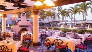 An outdoor dining area with tables and chairs, a central fire feature, and palm trees in the background. - Ocean front with new,updated interior.  Oversized balcony with BBQ. Perfect!! (Cabo San Lucas)