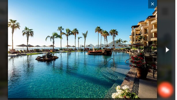 A resort pool with clear water, palm trees, and a building in the background. - Ocean front with new,updated interior.  Oversized balcony with BBQ. Perfect!! (Cabo San Lucas)
