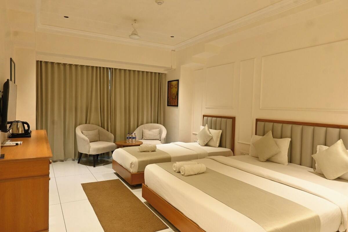 A hotel room with two single beds, a desk with a kettle, two armchairs, a coffee table, and a large window with curtains.