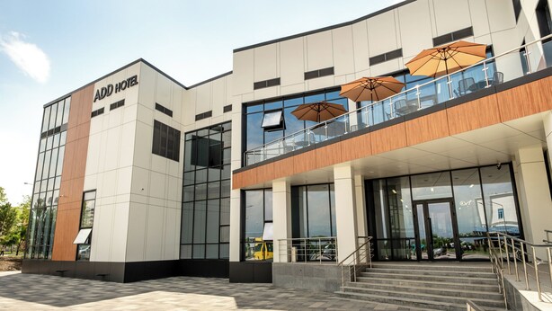 Un hôtel moderne, reconnaissable à son grand panneau « ADD HOTEL », à son entrée en verre, à son espace extérieur aménagé avec des parasols et à un escalier menant à l’entrée.