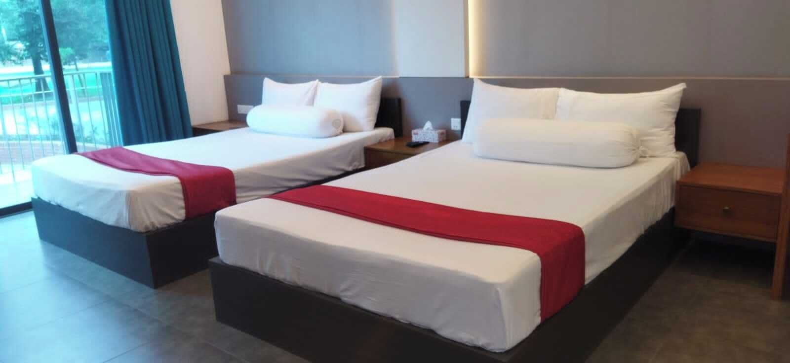A hotel room with two beds, each with white bedding and red throw blankets. The room has a modern design with a large window providing a view of the outdoors.