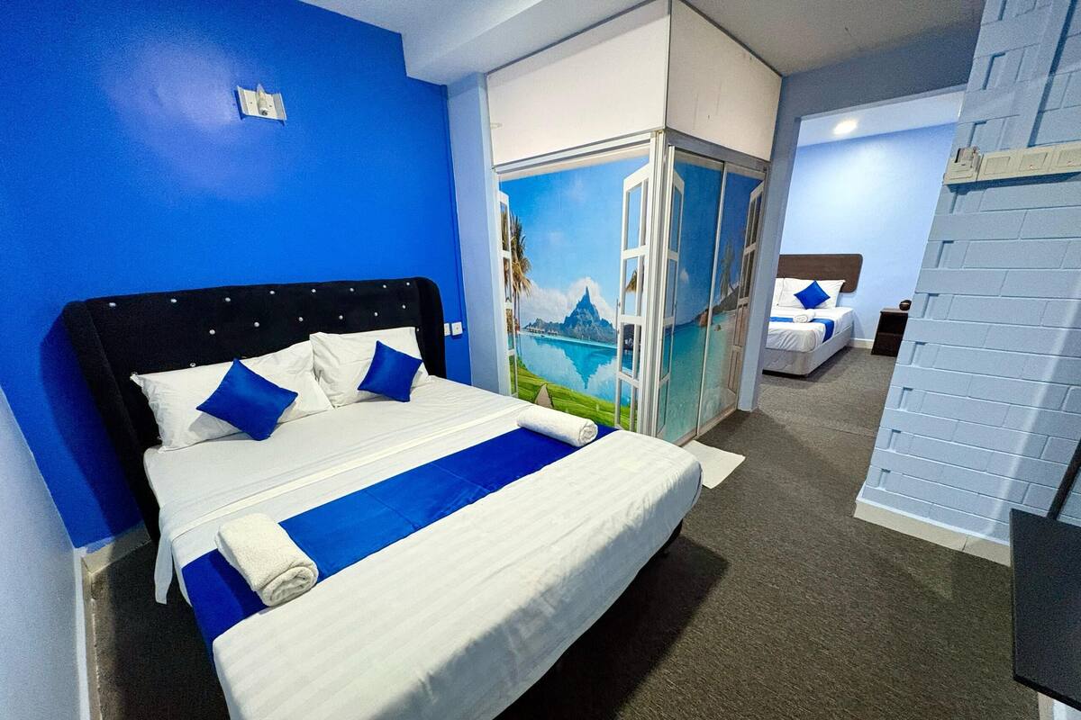 Una habitación de hotel con una cama grande, una pared azul decorativa, un mural de un paisaje y una cama separada al fondo.