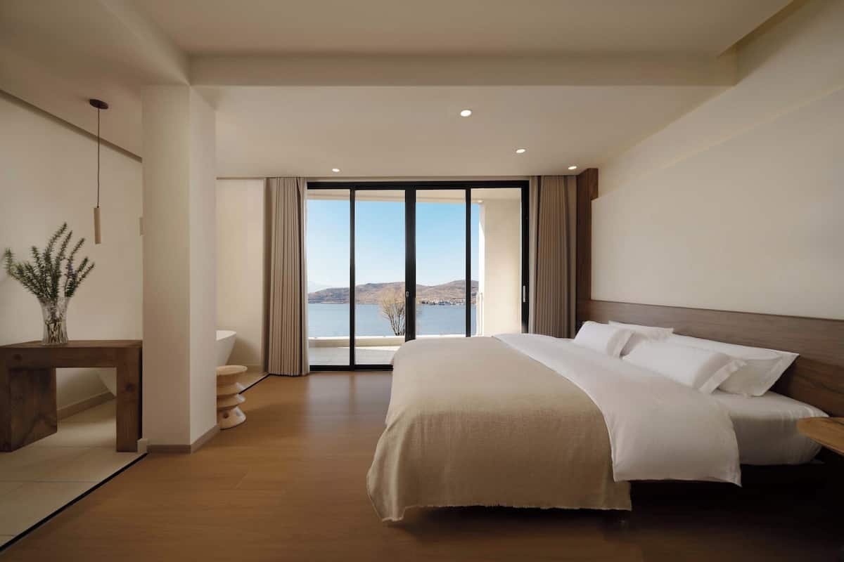 Une chambre d’hôtel moderne dotée d’un grand lit, d’une table de chevet en bois, d’un bureau et offrant une vue sur la mer grâce à de grandes fenêtres du sol au plafond.