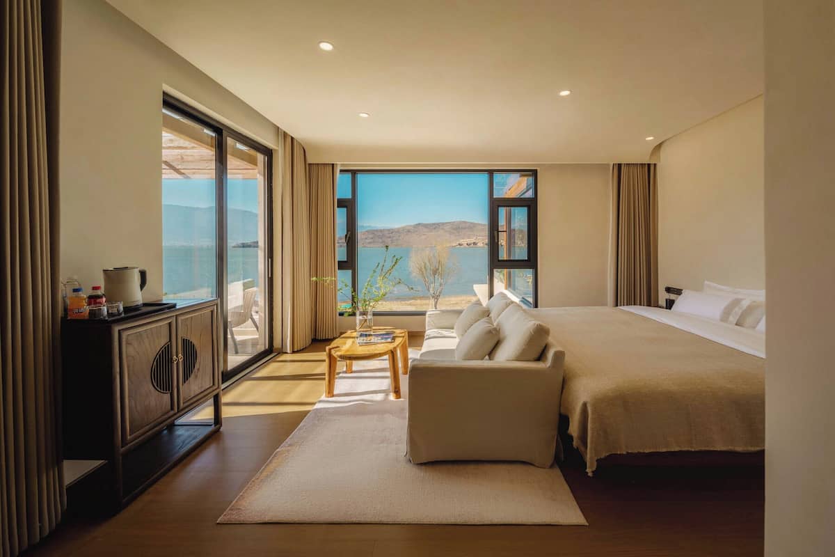 Une chambre d’hôtel moderne avec un grand lit, un canapé, une table basse en bois et une vue sur la mer grâce à de grandes fenêtres.
